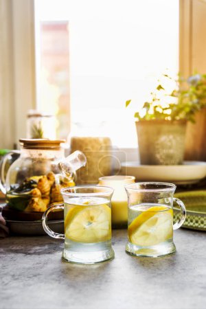 Téléchargez les photos : Boisson saine au citron et au gingembre dans des tasses à thé en verre à la table de cuisine avec des tasses, des bougies et des herbes au fond de la fenêtre avec de la lumière naturelle. Réchauffer les boissons chaudes aux agrumes. Vue de face. - en image libre de droit