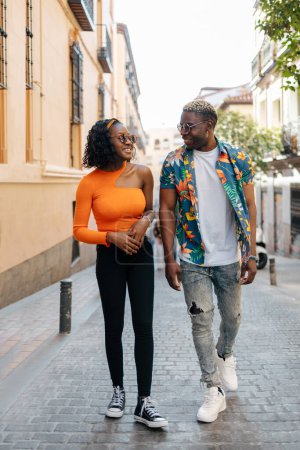 Téléchargez les photos : Jeune couple noir portant des lunettes de soleil et des vêtements colorés se regardant tout en se promenant dans la ville par une journée ensoleillée - en image libre de droit