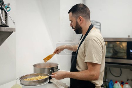 Téléchargez les photos : Jeune chef barbu vêtu d'un tablier de cuisine cuisinant des légumes dans une casserole attendant d'ajouter des pois chiches dans une cuisine bien équipée - en image libre de droit