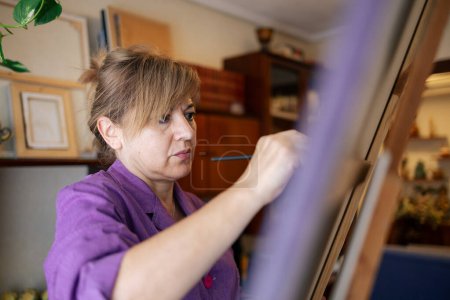 Photo for Adult woman with blonde hair dressed in a purple robe paints with brush on canvas in her home - Royalty Free Image