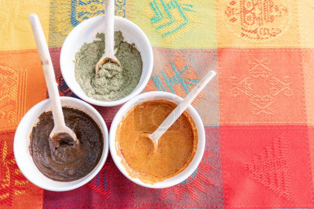 Téléchargez les photos : Trois bols avec de la sauce taupe rouge, verte et jaune avec une cuillère en bois sur le dessus d'une nappe traditionnelle colorée. Concept de cuisine mexicaine traditionnelle de Puebla et Oaxaca - en image libre de droit