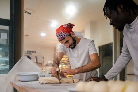 Téléchargez les photos : Jeune boulanger arabe vêtu d'un t-shirt blanc et d'un chapeau rouge coupant la pâte à l'omoplate avec un collègue noir qui le regarde dans une boulangerie - en image libre de droit