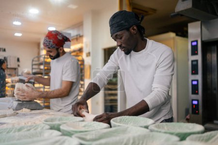 Junger schwarzer Bäcker mit weißem T-Shirt und schwarzem Hut knetet neben seinem Kollegen, der in einer Bäckerei Teig in Waage wiegt