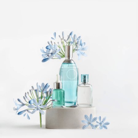 Beauty products in blue glass spray and pipette bottles standing on podium with flowers and petals at white background. Cosmetic setting with serum,perfume fragrance and toner. Front view