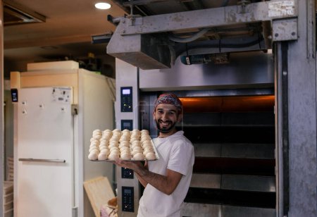 Photo for Portrait of a young arabic baker in a white t-shirt and red cap carrying a tray with a multitude of small dough balls smiling in a bakery with the oven in the background - Royalty Free Image