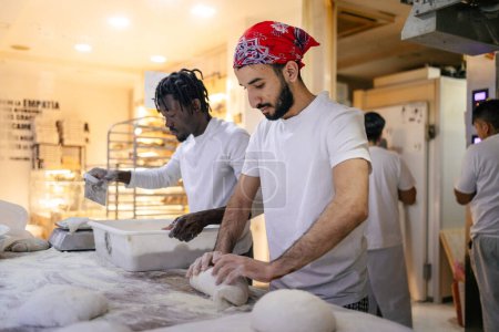 Foto de Panadero árabe en ropa blanca y gorra roja amasando para hacer pan con un compañero de trabajo negro a su lado cortando la masa en pedazos en una panadería - Imagen libre de derechos