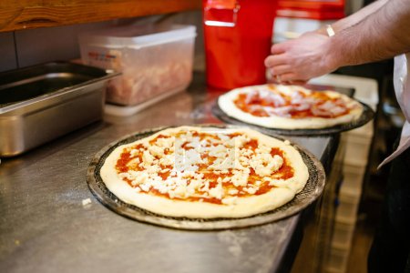 Téléchargez les photos : Portrait d'une pizza à la tomate et au fromage en préparation avant d'aller au four avec les mains du livreur de pizza en arrière-plan préparant une autre pizza - en image libre de droit