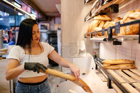 Téléchargez les photos : Jeune femme latina aux cheveux noirs et t-shirt blanc mettant un pain dans un sac avec un gant pour un client dans une boulangerie - en image libre de droit