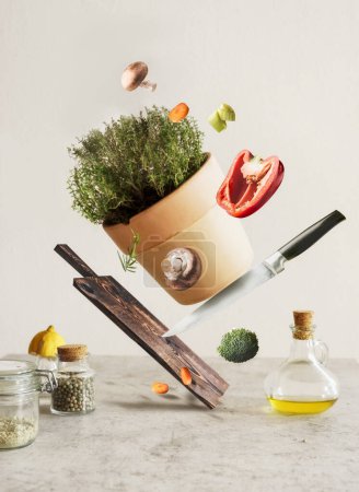 Foto de Concepto creativo de levitación de alimentos con tabla de cortar voladora, cuchillo, verduras, hierbas en macetas y utensilios en la mesa de cocina de hormigón gris. Cocinar en casa con ingredientes sabrosos y saludables - Imagen libre de derechos