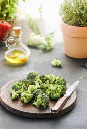 Photo for Broccoli on wooden cutting board with kitchen knife at grey kitchen table with herbs, oil and ingredients at background with natural lights - Royalty Free Image