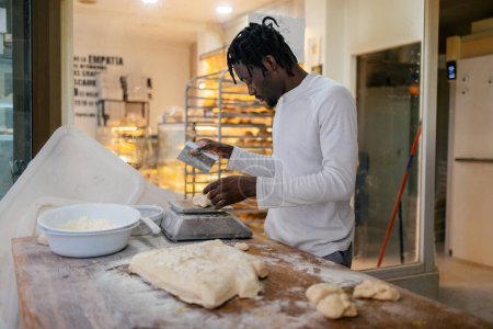 Foto de Joven panadero negro con rastas y ropa blanca cortando masa con una escápula y pesándola en una balanza en una panadería - Imagen libre de derechos