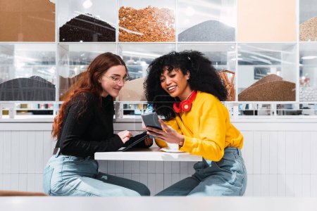 Téléchargez les photos : Vue latérale de joyeux jeunes femmes multiraciales meilleures amies en vêtements décontractés riant joyeusement tout en étant assis dans un café moderne avec des smartphones dans les mains - en image libre de droit