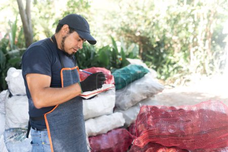 In an outdoor setting, a Hispanic worker meticulously records the weights of recycled glass bottle sacks, highlighting diligent monitoring within natural surroundings for sustainable practices
