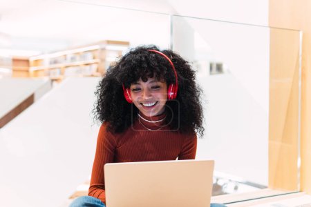 Foto de Negro rizado mujer alegre con auriculares mientras está sentado con el ordenador portátil en la biblioteca moderna - Imagen libre de derechos