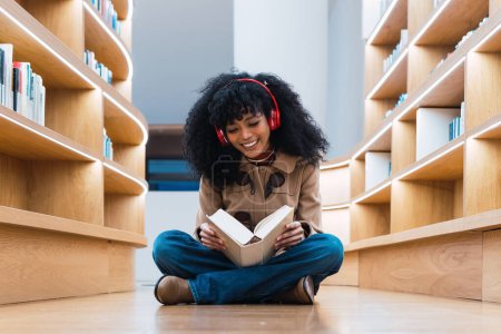Foto de Feliz estudiante afroamericana en auriculares leyendo libro mientras está sentada en el suelo en la biblioteca y disfrutando - Imagen libre de derechos