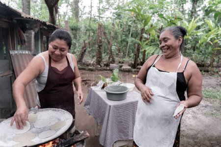 Deux femmes hispaniques adultes s'amusent à préparer des tortillas de maïs sur un poêle extérieur au Guatemala