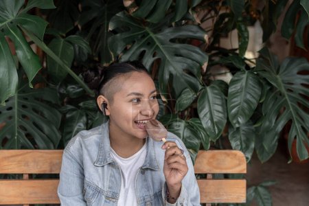 Téléchargez les photos : Une jeune femme hispanique déguste une glace tout en écoutant de la musique assise sur un banc avec des plantes en arrière-plan. Concept de snack sucré et de technologie - en image libre de droit