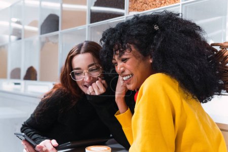 Photo for Side view of cheerful young multiracial female best friends in casual clothes laughing happily while sitting in modern cafe with smartphones in hands - Royalty Free Image