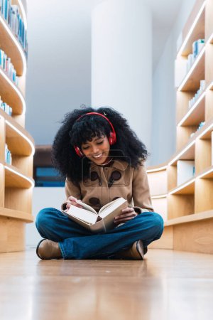 Foto de Feliz estudiante afroamericana en auriculares leyendo libro mientras está sentada en el suelo en la biblioteca y disfrutando - Imagen libre de derechos