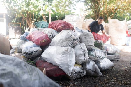 A Young Hispanic Woman stacks glass bottle sacks against a backdrop of a towering mountain of recycled heaps, contributing to sustainable efforts in recycling