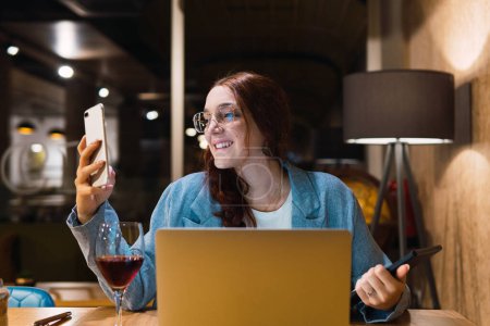 Foto de Sonriente joven inteligente en gafas de lectura y análisis de datos en el teléfono inteligente mientras está sentado en la mesa con gadgets y vino tinto durante el trabajo en el restaurante por la noche - Imagen libre de derechos