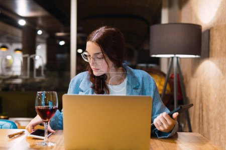 Photo for Smart young woman in glasses reading and analyzing data on smartphone while sitting at table with gadgets and red wine during work in restaurant in evening - Royalty Free Image