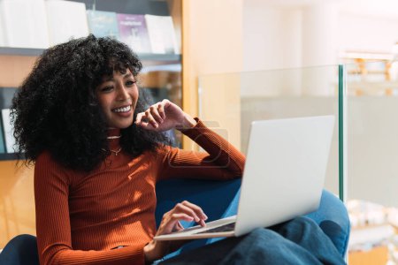 Téléchargez les photos : Femme afro-américaine satisfaite avec la coiffure afro assis dans le fauteuil dans la bibliothèque et en utilisant un ordinateur portable tout en travaillant sur le projet - en image libre de droit