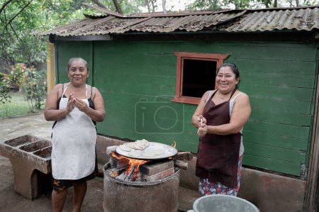 Two adult Hispanic women are smiling while shaping corn tortillas by hand and using an outdoor stove in Guatemala