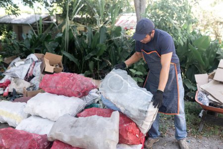 Téléchargez les photos : Un Hispanique empile un sac rempli de bouteilles en verre recyclé sur une grande pile de sacs similaires, s'engageant dans le processus de recyclage et contribuant à des pratiques durables - en image libre de droit