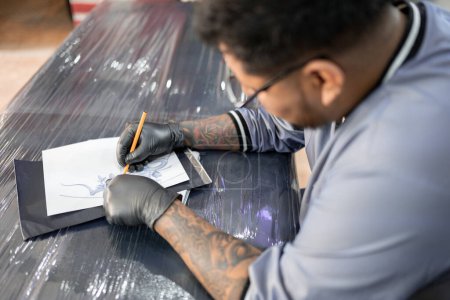 Téléchargez les photos : Un tatoueur dessine un pochoir sur sa civière de travail avant le tatouage. Concept d'inspiration d'artiste - en image libre de droit