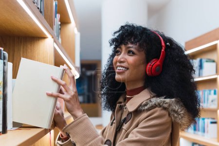 Téléchargez les photos : Vue latérale de dame afro-américaine positive écoutant de la musique et prenant le livre de l'étagère de la bibliothèque - en image libre de droit
