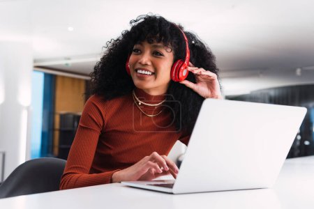 Photo for Content African American female in headphones using phone sitting at table with laptop in modern workspace - Royalty Free Image
