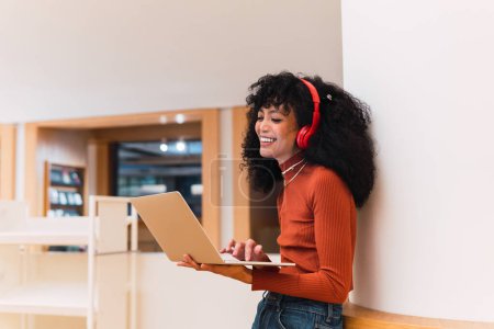 Foto de Negro rizado mujer alegre con auriculares mientras está de pie con el ordenador portátil en la biblioteca moderna apoyada en la pared - Imagen libre de derechos