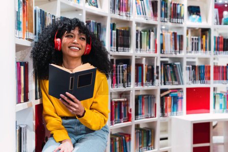 Foto de Elegante joven estudiante étnica feliz con pelo largo y rizado oscuro en traje de moda y auriculares en el cuello sonriendo mientras lee el libro en la biblioteca moderna - Imagen libre de derechos