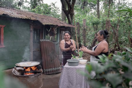 Two adult Hispanic women are smiling and shaping corn tortillas by hand and using an outdoor stove in Guatemala