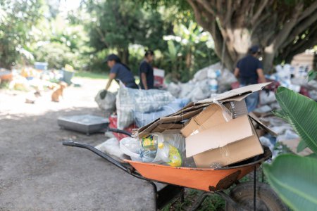 In the forefront stands a loaded wheelbarrow filled with plastics and cardboard, while workers blur in the background, working in recycling activities