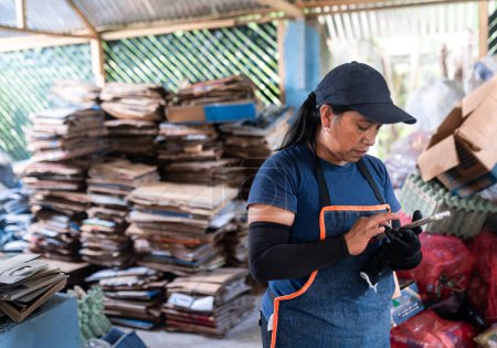 Eine erwachsene hispanische Frau dokumentiert auf ihrem Handy in einer Recyclinganlage fleißig Details zum Kartonstapeln