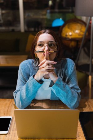 Foto de Alto ángulo de la joven mujer de negocios en gafas que sostienen la pluma bajo la nariz y tocar los labios mientras está sentado en la mesa con el ordenador portátil y reflexionar sobre los datos en la noche - Imagen libre de derechos