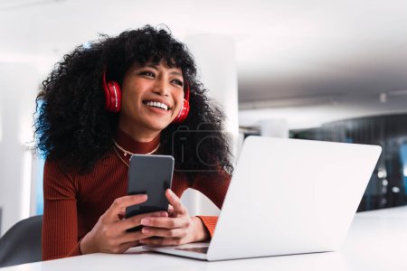 Content African American female in headphones using phone sitting at table with laptop in modern workspace