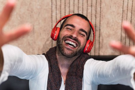 Téléchargez les photos : Grand angle de mélomane masculin joyeux en chemise blanche souriant et tendre la main à la caméra tout en écoutant de la musique dans des écouteurs sans fil - en image libre de droit