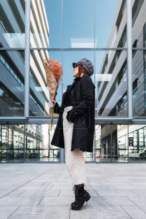 Photo for Side view of young woman in stylish outerwear with cap and glasses holding orange flowers against modern building with glass walls on city street - Royalty Free Image