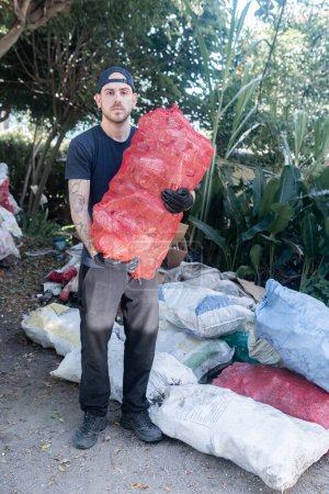 Foto de Un joven caucásico sostiene una bolsa llena de botellas de vidrio reciclado en una planta de reciclaje, mostrando una expresión seria mientras trabaja en el reciclaje en un entorno industrial - Imagen libre de derechos