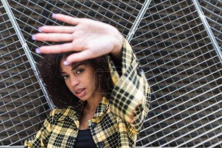 Young trendy ethnic female in checkered jacket demonstrating control gesture while looking at camera in daylight