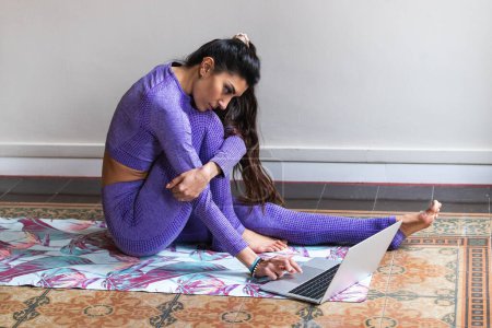 Téléchargez les photos : Vue latérale de la jeune dame hispanique positive avec de longs cheveux noirs assis sur le tapis et l'ordinateur portable de navigation tout en pratiquant le yoga à la maison - en image libre de droit