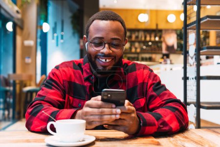 Téléchargez les photos : Homme barbu afro-américain souriant en chemise à carreaux rouges messages sur les écouteurs d'esprit de téléphone mobile tout en buvant du café dans le café et en regardant l'écran - en image libre de droit