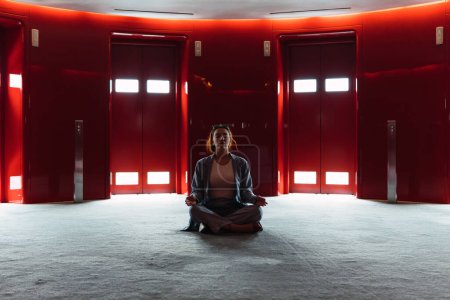 Téléchargez les photos : Corps entier de femme paisible assis dans la pose du lotus tout en méditant dans le hall spacieux avec des ascenseurs rouges avec des lumières éclatantes - en image libre de droit