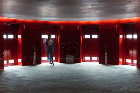 Foto de Mujer anónima distante en elegante desgaste de pie cerca de modernos ascensores rojos con luces brillantes en el amplio vestíbulo con poca luz - Imagen libre de derechos