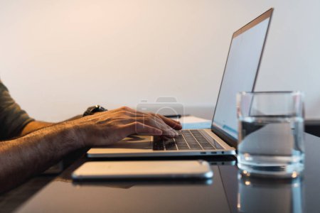 Téléchargez les photos : Crop anonyme travailleur masculin naviguant netbook portable moderne tout en étant assis à table avec smartphone dans la pièce lumineuse pendant le processus de travail - en image libre de droit