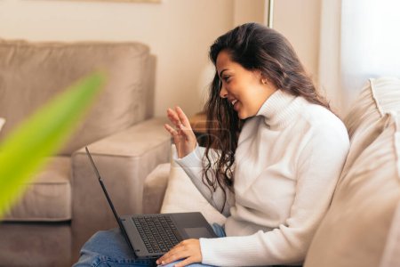 Téléchargez les photos : Une jeune femme parle lors d'un appel vidéo avec un ordinateur portable à la maison, souriant et regardant engagé dans la conversation. - en image libre de droit