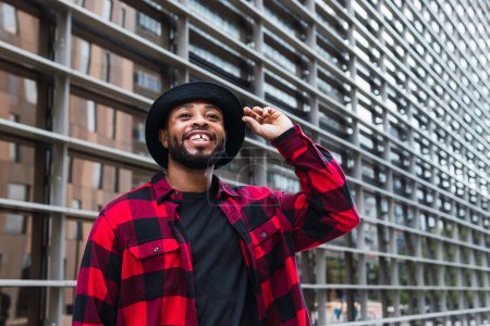 Foto de Varón afroamericano barbudo positivo con camisa a cuadros roja y sombrero negro de pie cerca de la pared de vidrio en la calle de la ciudad y mirando hacia otro lado - Imagen libre de derechos